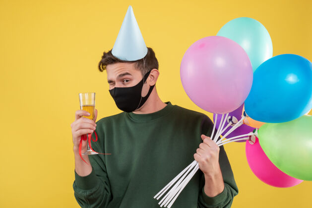 男人正面图：戴着派对帽 戴着黑色面具 手持酒杯和黄色气球的年轻人年轻人酒黑
