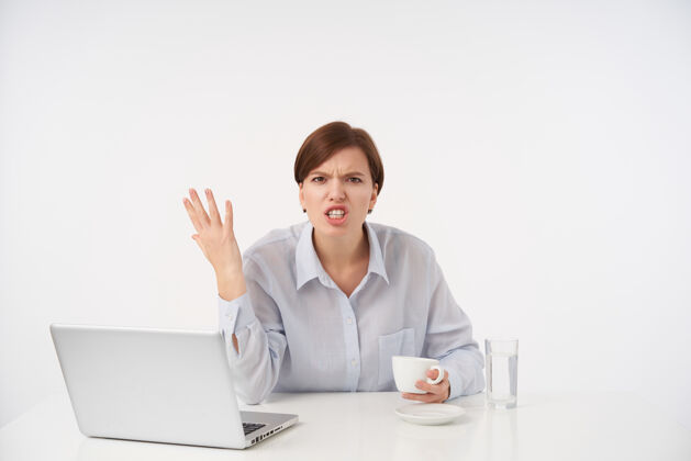 现代恼怒的棕色头发的年轻女人 自然化妆 情绪化地举起她的手 愤怒地看着 有压力的一天 坐在白色笔记本电脑员工休闲