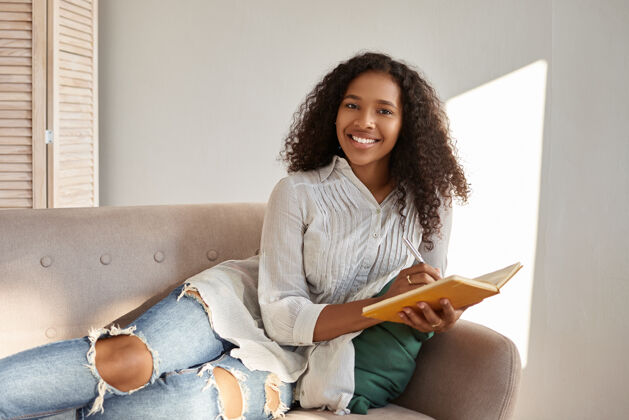 现代人 生活方式 休闲 爱好和休息可爱迷人的年轻黑皮肤女人 非洲式发型 在舒适的灰色沙发上放松 微笑着 在日记里写下目标和计划笔记本笔记肖像