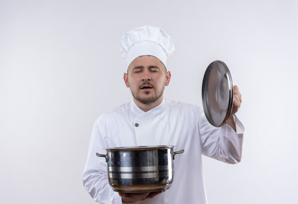 年轻高兴的年轻帅哥厨师穿着厨师制服拿着锅和锅盖闭上眼睛隔离在白色空间持有盖子锅