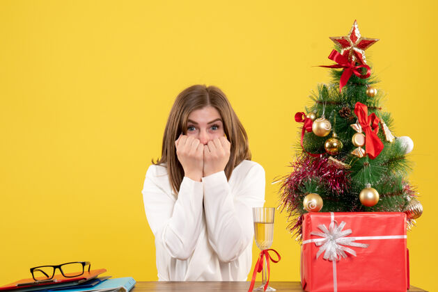 坐正面图：女医生坐在黄色背景的桌子前 放着圣诞树和礼品盒前面圣诞节笔记本电脑