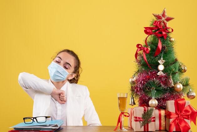 情感正面图女医生戴着黄色背景的无菌口罩坐在圣诞树和礼盒里看面具人
