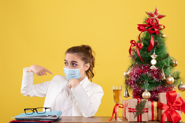 商务正面图女医生戴着黄色背景的无菌口罩坐在圣诞树和礼盒里职业面具人