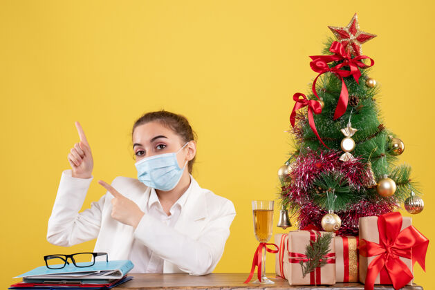 微笑正面图女医生戴着黄色背景的无菌口罩坐在圣诞树和礼盒里情绪前面肖像
