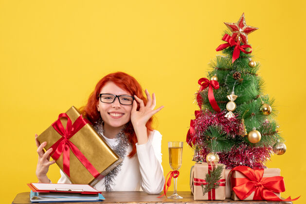 漂亮前视图：女医生拿着圣诞礼物和黄色背景上的圣诞树坐着圣诞树女性情绪