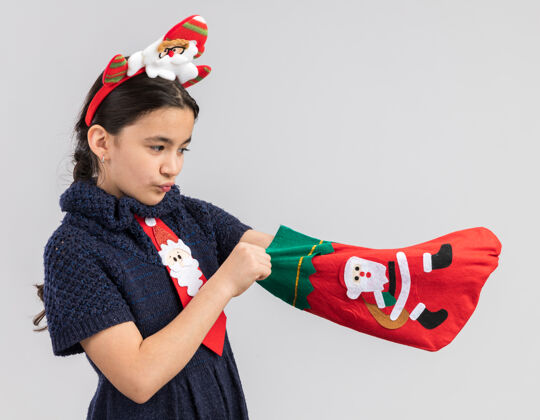 小小女孩穿着针织连衣裙 头上系着一条红色领带 头上戴着一个有趣的圣诞边 手里拿着圣诞长袜 看起来很好奇新年领带头巾