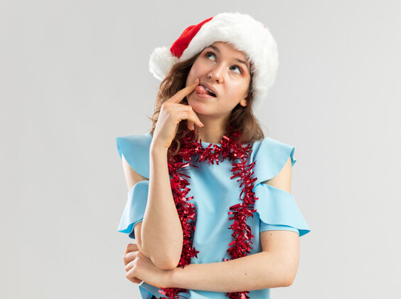 帽子身穿蓝色上衣 戴着圣诞帽 脖子上戴着金属丝的年轻女子 疑惑地抬起头来圣诞老人向上年