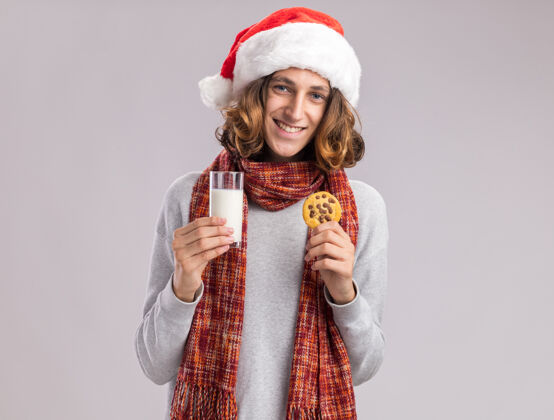 抱着快乐的年轻人戴着圣诞老人的帽子 脖子上围着暖和的围巾 手里拿着一杯牛奶和饼干 看上去笑容满面看着男人新年