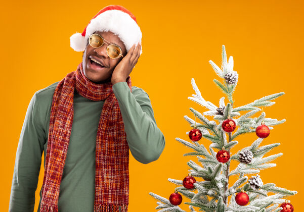 男人戴着圣诞帽 脖子上围着围巾的非裔美国人站在橙色墙壁上的圣诞树旁 开心而积极地看着一边非洲圣诞节脖子
