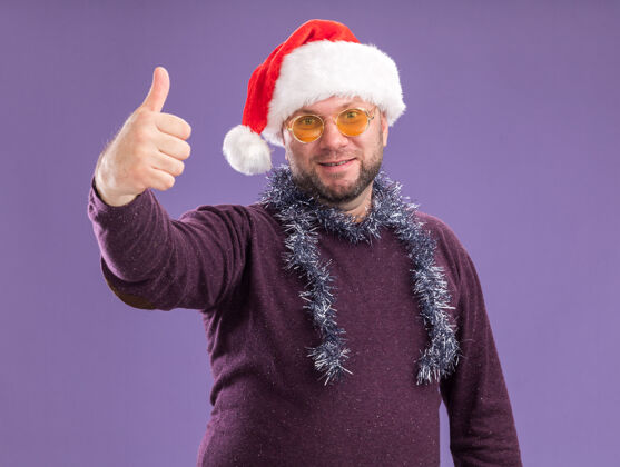花环一个戴着圣诞老人帽 脖子上戴着金箔花环 戴着眼镜的中年男人孤零零地站在紫色的墙上高兴展示帽子