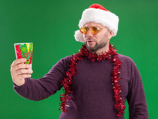中年困惑的中年男子戴着圣诞帽 脖子上戴着金属丝花环 戴着眼镜 伸出塑料圣诞杯帽子脖子周围