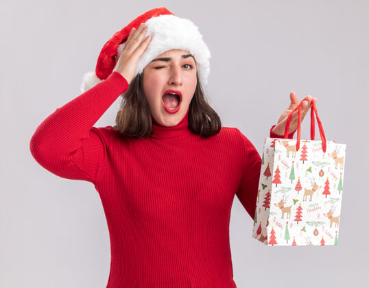 站着身穿红色毛衣 头戴圣诞帽的小女孩手拿彩色纸袋 手里拿着圣诞礼物 惊讶地站在白色的墙上圣诞老人毛衣圣诞节