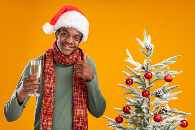 圣诞节戴着圣诞帽 围着围巾的非洲裔美国人 手拿香槟酒杯 在橙色背景下的圣诞树旁愉快地竖起大拇指展示微笑玻璃杯