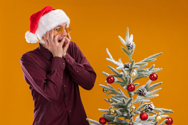 近兴奋的年轻金发男子戴着圣诞帽和眼镜站在装饰过的圣诞树旁 手放在脸上 孤立地看着橙色背景圣诞树圣诞看