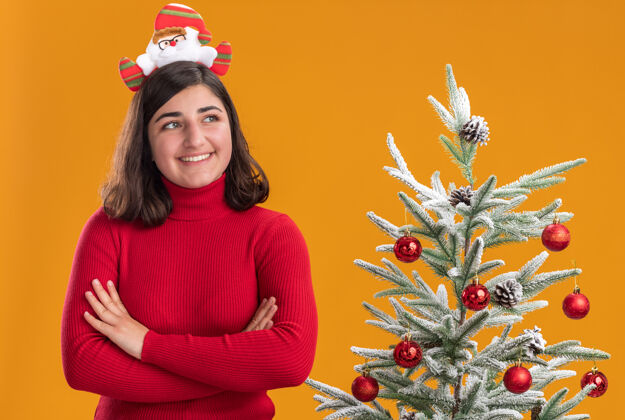 搞笑快乐的小女孩穿着圣诞毛衣戴着滑稽的头带 脸上挂着微笑 站在橙色背景下的圣诞树旁交叉圣诞节微笑