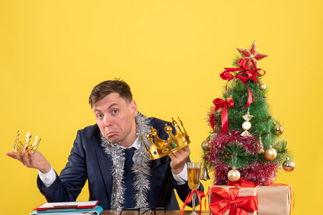 花商务人士双手捧着皇冠坐在圣诞树旁的桌子前 黄色的礼物抱着男人黄色
