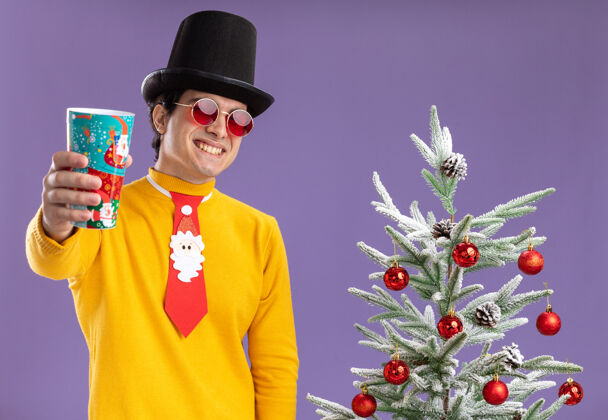 展示穿着黄色高领毛衣戴着眼镜戴着黑色帽子和滑稽领带的快乐的年轻人展示着五颜六色的纸杯微笑着快乐地站在紫色背景下的圣诞树旁搞笑树男人