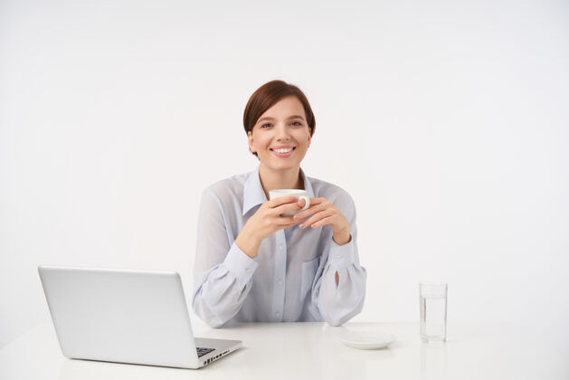 工作迷人的年轻棕色眼睛短发女士举起手拿着一杯咖啡 愉快地微笑着 带着笔记本电脑在现代办公室工作女士优雅办公室