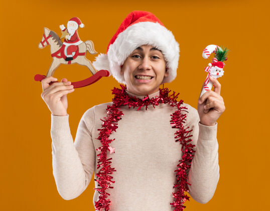 微笑微笑的年轻人戴着圣诞帽 脖子上戴着花环 手里拿着黄色背景上孤立着的糖果圣诞玩具花环圣诞节年轻人