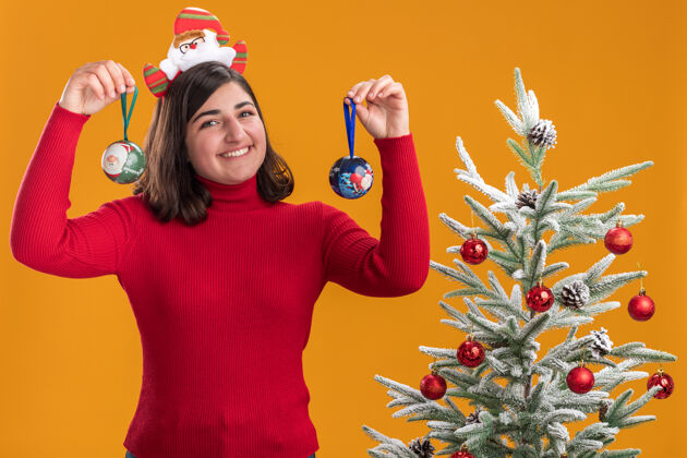 舞会穿着圣诞毛衣的快乐小女孩戴着滑稽的头带 手拿圣诞球 脸上带着微笑 站在橙色墙上的圣诞树旁毛衣站着头带