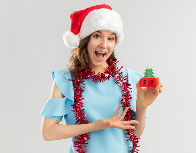 新年身穿蓝色上衣 戴着圣诞帽 脖子上戴着金属丝的年轻女子 手挽着圣诞礼物玩具立方体 快乐而兴奋手臂帽子手