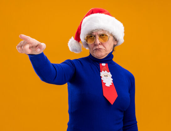 橙色戴着太阳眼镜 戴着圣诞帽 系着圣诞领带的恼怒的老妇人看着并指着在橙色背景上有复制空间的孤立的一面圣诞老人领带背景