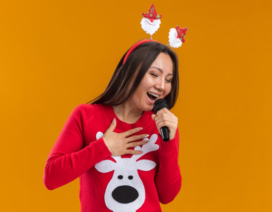 快乐快乐的年轻亚洲女孩戴着圣诞发箍和毛衣在橙色墙上的麦克风上唱歌女人麦克风年轻