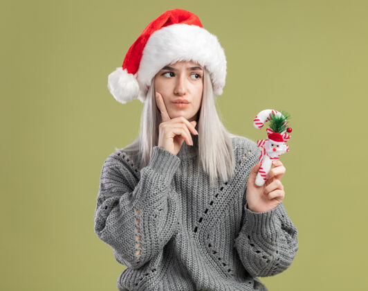金发年轻的金发女人穿着冬天的毛衣 戴着圣诞帽 手里拿着圣诞糖果手杖 困惑地看着一边圣诞快乐拿着一旁