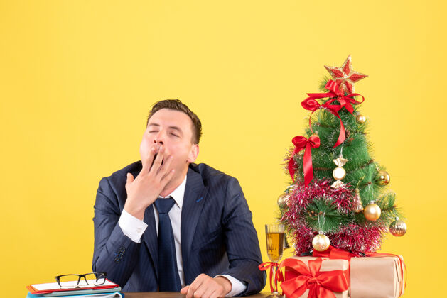 瞌睡男人睡意朦胧的男人正坐在圣诞树旁的桌子旁打哈欠 黄色的礼物坐着商人职业