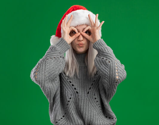 年轻年轻的金发女郎穿着冬季毛衣 戴着圣诞帽 透过手指做着双眼的姿势 站在绿色的背景下 快乐而积极圣诞金发制作