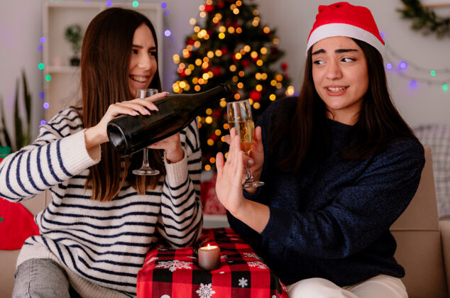 停戴着圣诞帽的漂亮女孩拿着一杯香槟 向坐在扶手椅上的朋友打着停车牌 手里拿着一瓶香槟 在家里享受圣诞时光坐着圣诞老人家