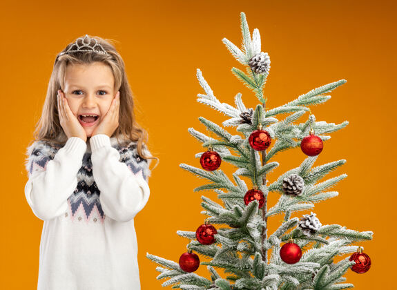 花环兴奋的小女孩站在圣诞树旁 戴着头饰 脖子上戴着花环 手放在橘色的墙上孤立的脸颊上头饰放兴奋