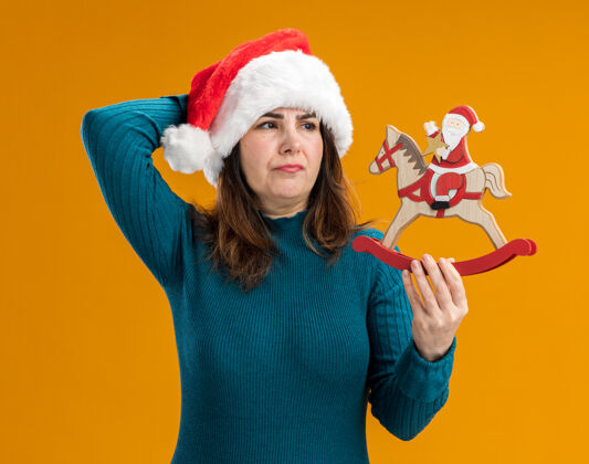 圣诞老人未出租的成年白人妇女拿着圣诞老人的帽子 看着圣诞老人在摇马装饰孤立的橙色背景与复制空间举行成人未租赁