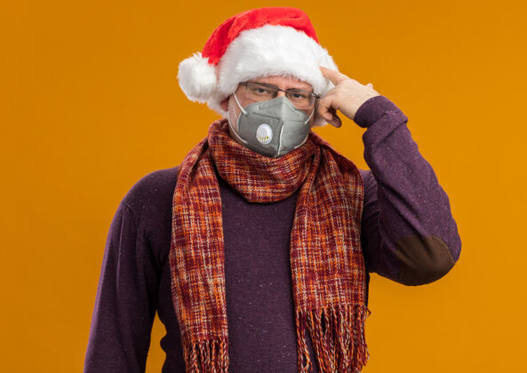面具受够了戴着防护面具眼镜戴着圣诞老人帽子脖子上围着围巾看着摄像机在橙色背景下做动作手势眼镜