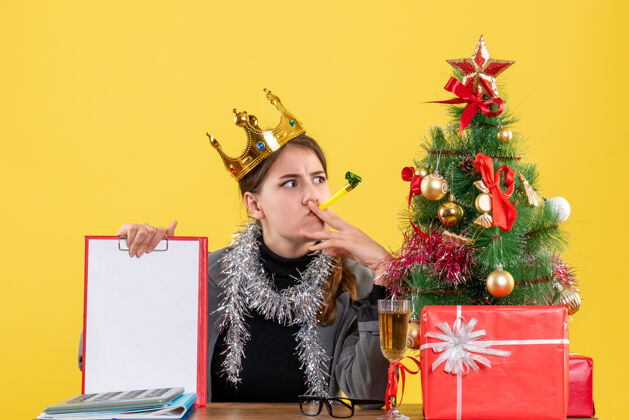 坐着正面图贴心的戴着皇冠的女孩坐在桌子旁 手里拿着圣诞树和鸡尾酒礼物皇冠圣诞节装饰
