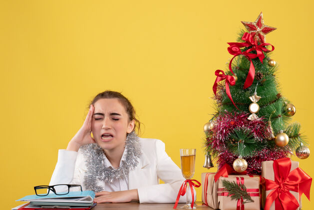 情绪正面图：坐在桌子后面的女医生 带着圣诞树和礼盒 在黄色的背景下 她感到悲伤悲伤感觉女医生