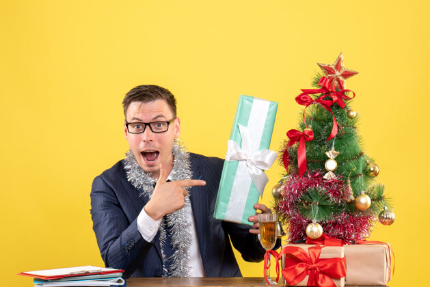 导演前视图是一个好奇的人指着坐在圣诞树旁的桌子上的礼物和黄色的礼物笔记本电脑礼物商人