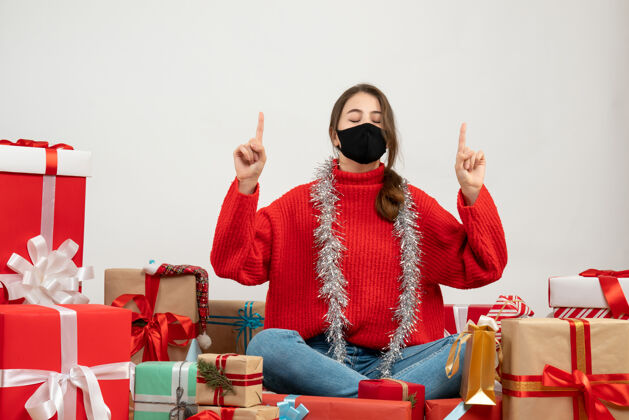 周围拿着红色毛衣的年轻女孩 手指朝上 围坐在礼物旁边 白色的面具上戴着黑色的面具男人面具圣诞节