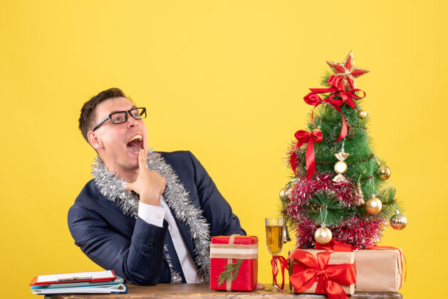 男人正面图：快乐的男人张嘴坐在圣诞树旁的桌子旁 黄色的礼物圣诞桌子商人