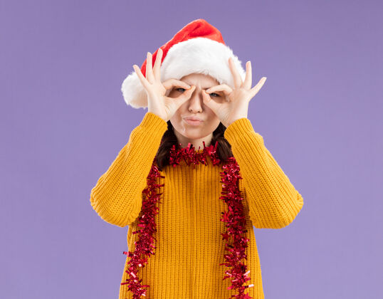 圣诞老人令人印象深刻的年轻斯拉夫女孩戴着圣诞帽 脖子上戴着花环 通过手指看着相机 在紫色背景上与复制空间隔离年轻帽子手指