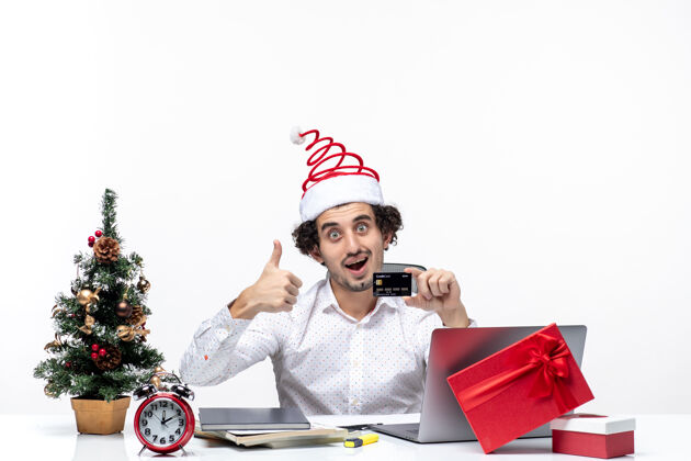 手势戴着圣诞老人帽子 拿着银行卡的商人在办公室里看着 做着“ok”的手势抱着圣诞老人帽子