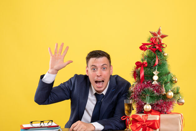 男前视图的快乐男子招呼某人坐在圣诞树附近的桌子上 并在黄色的礼物某人男人树