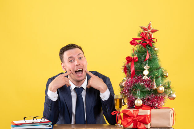 快乐圣诞树和黄色礼物旁的快乐男人坐在桌子前的正面视图圣诞坐着商人