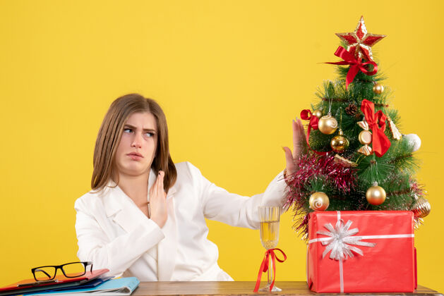 微笑正面图：女医生坐在黄色桌子上的桌子前 桌上放着圣诞树和礼品盒漂亮成人微笑