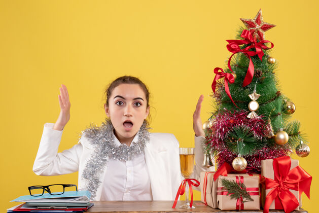 人物正面图：女医生坐在黄色背景的桌子后面 还有圣诞树和礼品盒女性圣诞节微笑