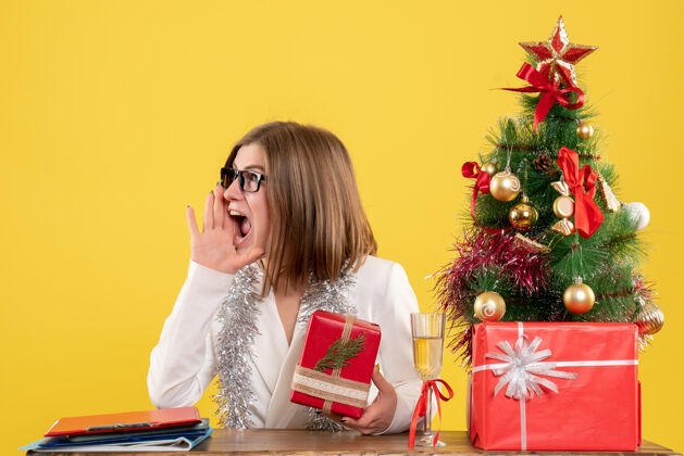 前面正面图：女医生坐在桌子前 拿着礼物 黄色背景上的圣诞树和礼盒办公室医生情感