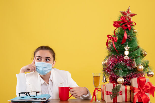 人正面图女医生戴着防护面具坐在黄色背景上 戴着圣诞树和礼品盒情感健康面具