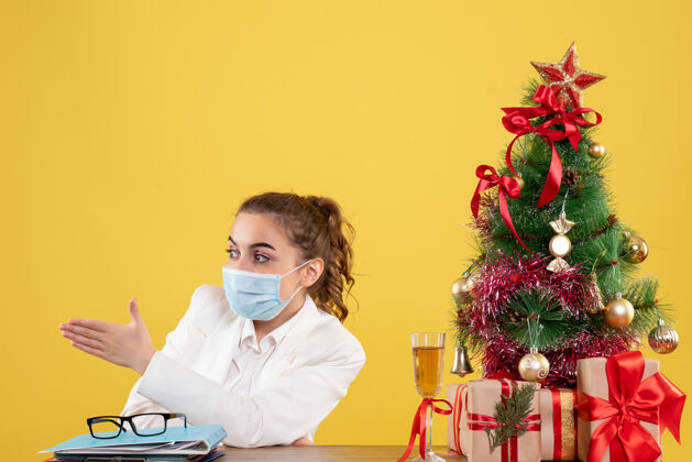 实验室外套正面图女医生戴着防护面具坐在黄色背景上 戴着圣诞树和礼盒面具人医生
