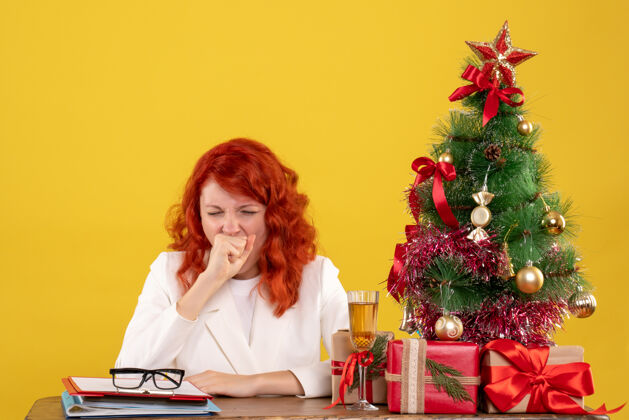 坐着前视图：女医生坐在桌子后面 黄色背景上打着哈欠的圣诞礼物圣诞节桌子微笑