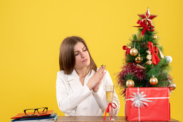 桌子正面图：女医生坐在黄色背景的桌子前 放着圣诞树和礼品盒微笑微笑肖像